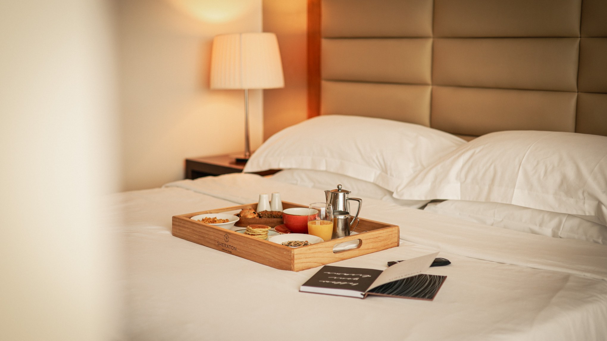 Breakfast tray with drinks and snacks on a hotel bed with white linens.