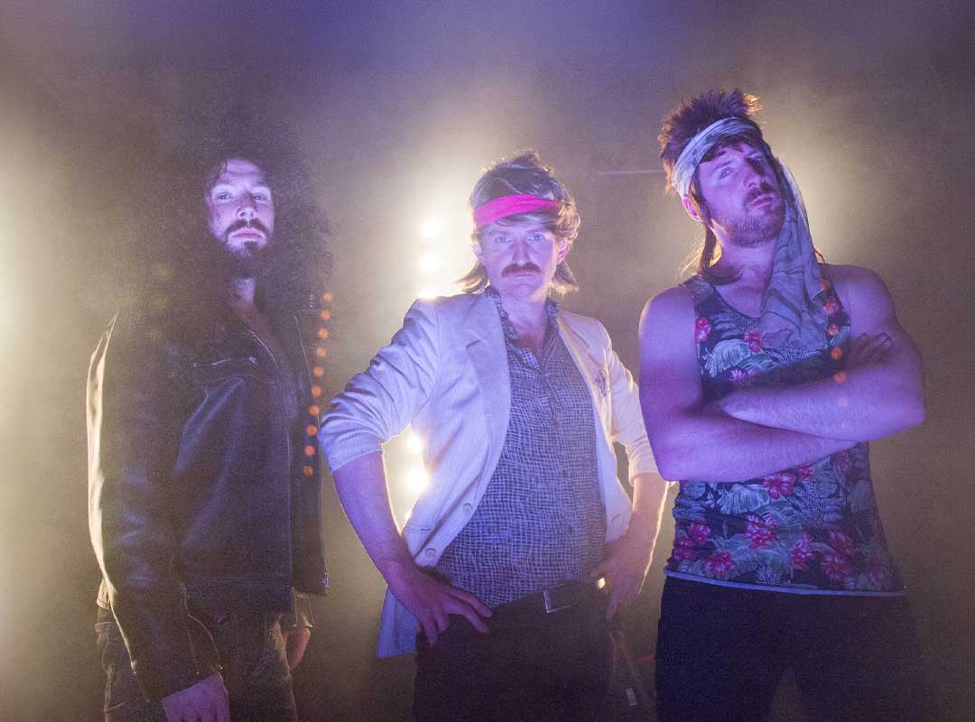 Three men in retro attire pose with colorful lighting and fog.