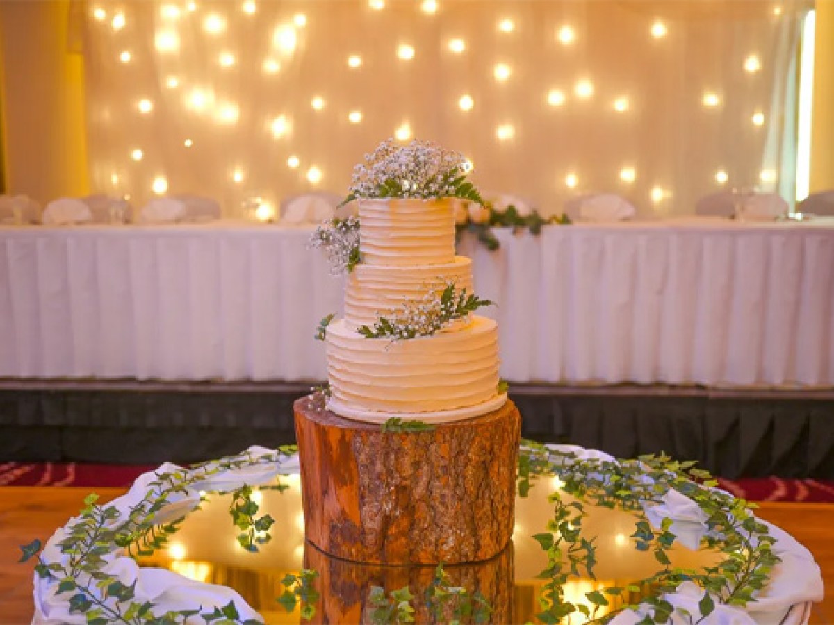 Three-tier wedding cake on a wooden stand, with greenery, in front of a lit backdrop during Sheraton Athlone Hotel wedding.